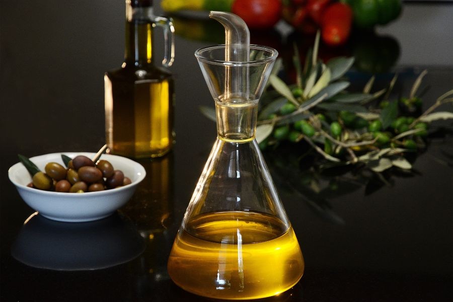 Önde yağdanlık içinde natürel sızma zeytinyağı arakda kasede yeşil zeytin ve masada zeytin dalları ve yaprakları