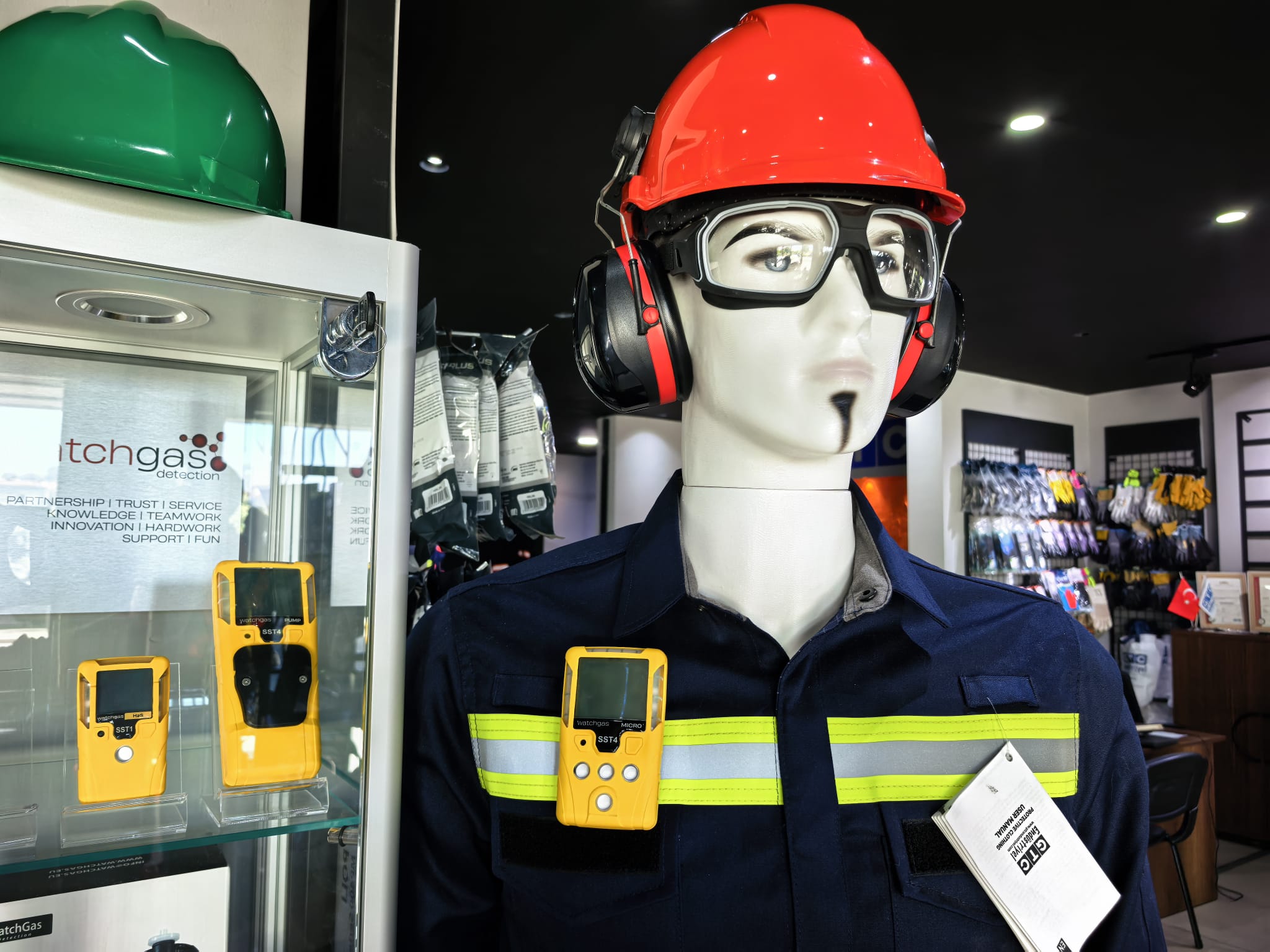 Baret, kulaklık, koruyucu gözlük ve reflektif iş elbisesiyle giydirilmiş manken; yanında WatchGas gaz ölçer cihazları sergileniyor