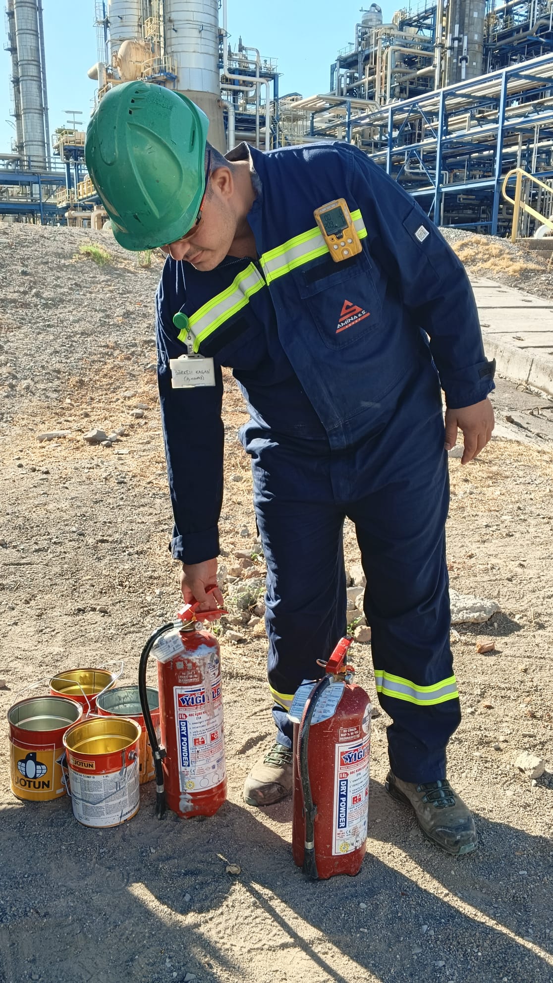 Petrokimya tesisinde koyu mavi tulum ve baretle çalışan personel, yangın söndürücü ve ekipman kontrolü yapıyor